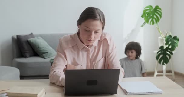 Młoda, skupiona kobieta pracująca zdalnie na laptopie. Mały uroczy syn podchodzi i przytula ją szczęśliwie. — Wideo stockowe