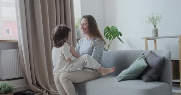 Delicioso, brincalhão, filho animado correndo, brincando, pulando e abraçando a jovem mãe reclinada no sofá. — Vídeo de Stock