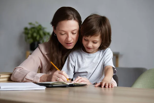 Usmívající se mladá máma pomáhá roztomilé syn dítě s domácím úkolem a cvičení. Domácí výuka — Stock fotografie