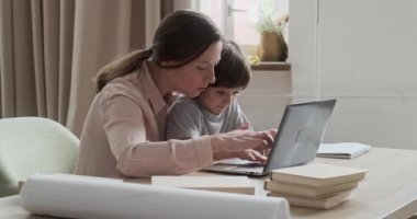 Neşeli anaokulu çocuğu bilgisayarla oynuyor ve oynaşıyor, ve anne genç kadın ders vermeye çalışıyor