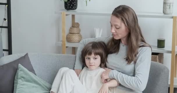 Cuidar madre calmar y abrazar poco adorable hijo sentado juntos en el sofá en el interior de casa — Vídeos de Stock