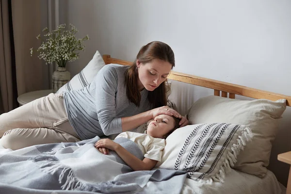 Moeder bezorgd zoon gezondheidscontrole warm voorhoofd liggend op bed kussen. Pijn in het hoofd van de kleuter — Stockfoto