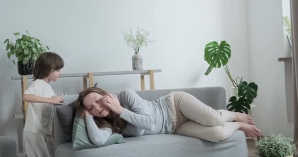 Mujer cansada tumbada en el sofá relajante comodidad interior del hogar. Lindo niño pequeño corriendo divertirse — Vídeos de Stock