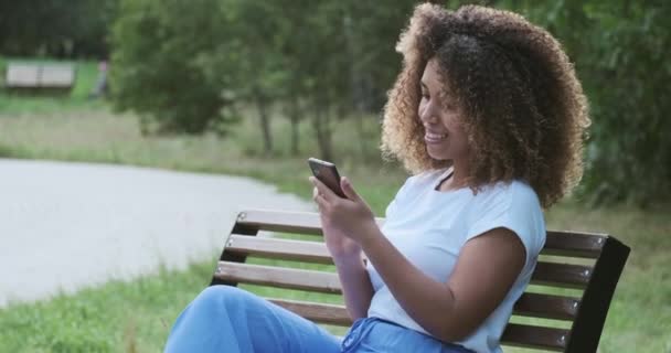 Ung svart afrikansk kvinna entreprenör i casuals sitter på bänken och talar via smartphone i parken. — Stockvideo