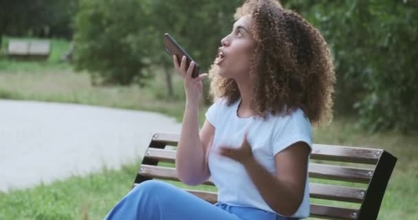Молодая черная женщина записывает аудиосообщение другу с помощью смартфона, сидящего на скамейке в парке. — стоковое видео