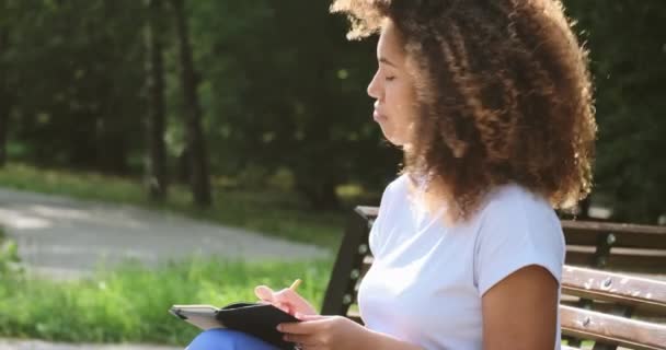 Jovem negra cuidadosa com cabelo encaracolado olha ao redor e escreve em um caderno de papel — Vídeo de Stock