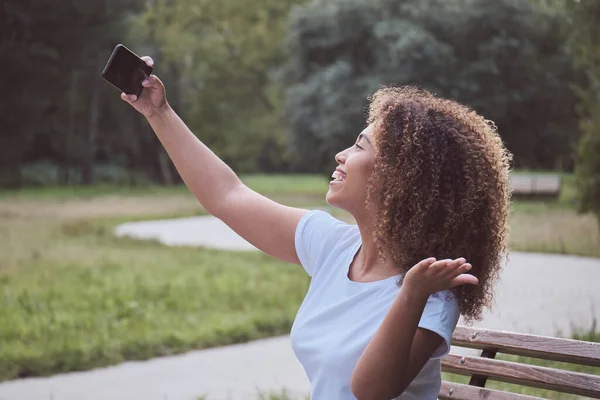 Rindo jovem pele preta mulher moderna posando tomando selfie usar smartphone sentado no parque de banco Imagens Royalty-Free