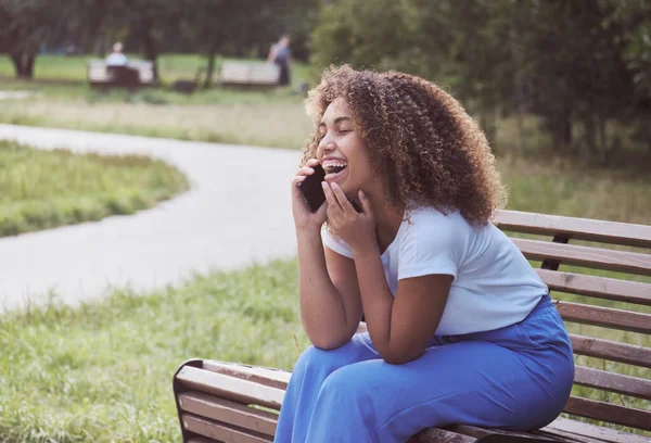 Jovem mulher negra africana empresária em casuals sentado no banco e falando sobre smartphone no parque. Fotografias De Stock Royalty-Free