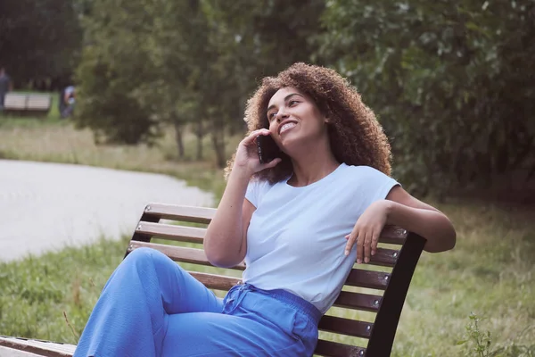 Jovem mulher negra africana empresária em casuals sentado no banco e falando sobre smartphone no parque. Imagens Royalty-Free