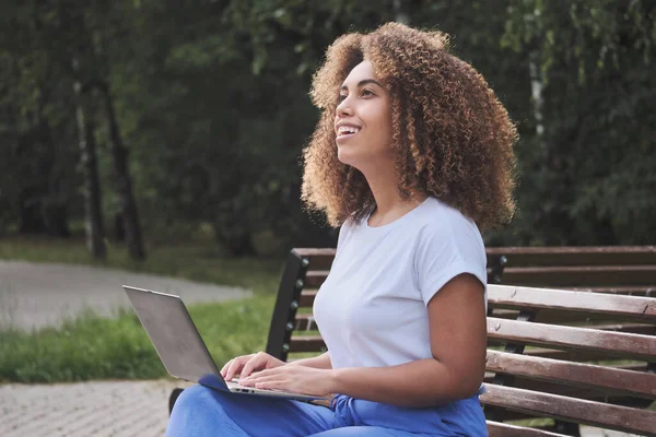 Smiling African modern woman with curly hair thinking working remotely online use laptop at park Stock Image