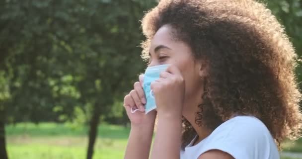 African curly woman trying medical face protective mask sitting on bench at summer forest park — Stock Video