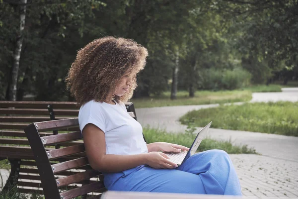 Modern African American woman focused remotely working use laptop sitting on bench at summer park Stock Photo