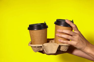 Sarı arka planda yakın plan, kadınların elleri bir stantta kahve ile iki kağıt bardak tutuyor. Seninle kahve ya da çay. Bir fiyatına iki terfi, sarı arka planda kahve teslimatı.