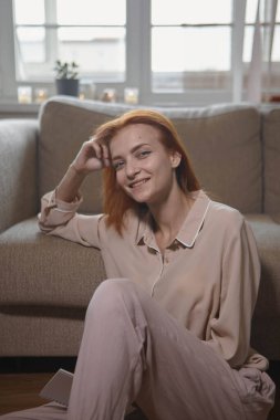 Beyaz, güzel, kızıl saçlı bir kadın odada koltuğun yanında oturuyor ve kadın kameraya bakıyor, gülümsüyor ve gülüyor. Neşe, sıcaklık, mutluluk..