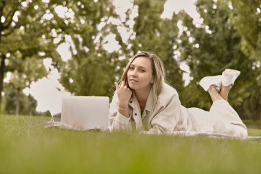 Kadın parkta dizüstü bilgisayar kullanarak yatıyor. Gülümseyen genç kız çimlerin üzerinde dizüstü bilgisayar kullanıyor..