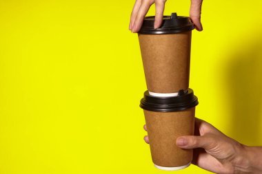 Sarı arka planda yakın plan, kadınların elleri bir stantta kahve ile iki kağıt bardak tutuyor. Seninle kahve ya da çay. Bir fiyatına iki terfi, sarı arka planda kahve teslimatı.