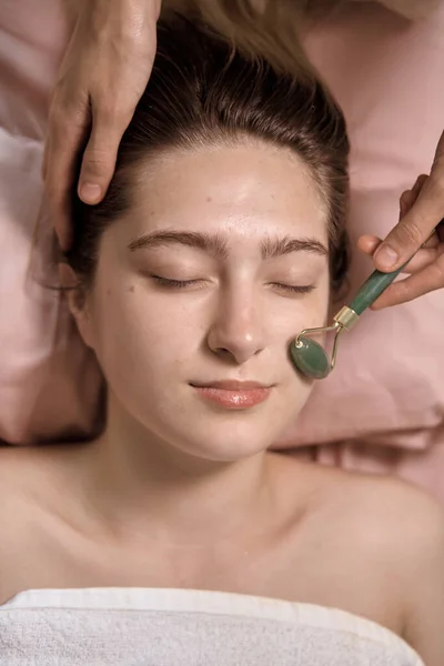 Young beautiful woman is getting a relaxing face Gua Sha Massage in wellness center. Masseur doing massage the head of an young beautiful girl woman in the spa salon close up macro