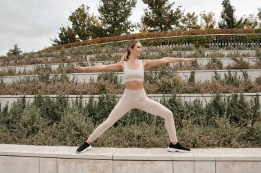 Orta yaşlı sarışın, beyaz kadın sporcu bej bluz giymiş ve tayt giymiş şehir parkında yoga asanası yapıyor. Ahenk ve barış. Açık havada yoga dersleri..