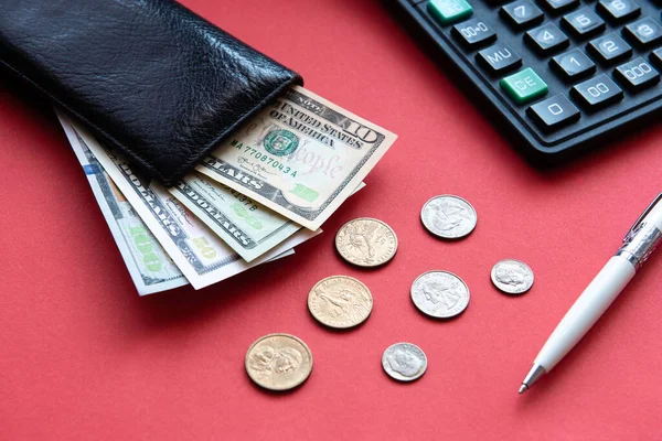 Kırmızı zemin üzerinde dolar banknotları, bozuk paralar, kalem ve hesap makinesi olan siyah deri bir cüzdan..