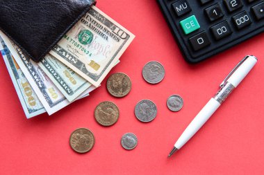Kırmızı zemin üzerinde dolar banknotları, bozuk paralar, kalem ve hesap makinesi olan siyah deri bir cüzdan..