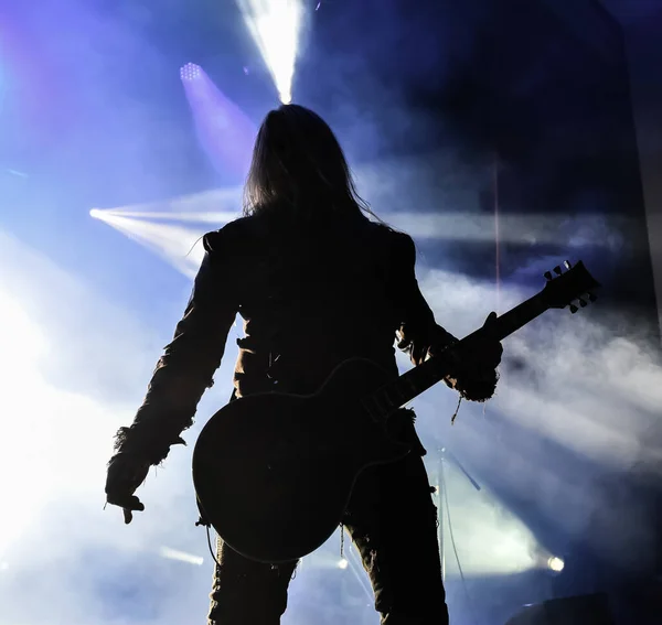 Sisteki bir konserde bir vokalist ve gitaristin silüeti. Karanlık arka plan, duman, konser ışıkları.