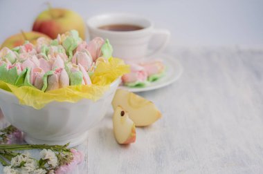 Beyaz bir tabakta lale şeklinde marşmelovların arka planında bulanık bir görüntü, elma dilimleri ve arka planda bir fincan çay..