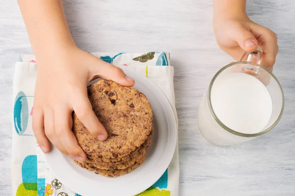 子供はミルクのカップで軽いテーブルの上のプレートから巨大なチョコレートチップクッキーを取ります — ストック写真