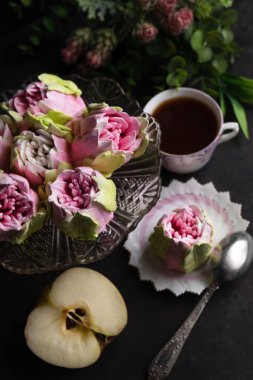 Çiçek şeklinde bulanık bir şekerlemenin görüntüsü, bir fincan kahve, siyah arka planda bir elma ve çiçekler. Hayat devam ediyor. Romantik bir geçmişi var. Yukarıdan bak..