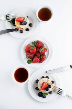 Beyaz masada çay fincanları, bir tabak çilek ve bir tabak gözleme ve meyve ve çatal bıçak takımı var. Çay partisi konsepti.