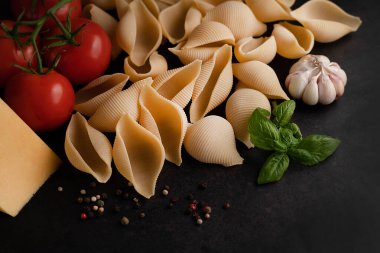 On a dark background, a blurred image of conchiglioni pasta, tomatoes, garlic, basil sprig and cheese.