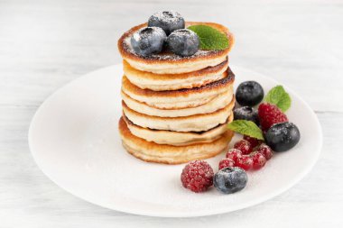 Pancakes with powdered sugar, blueberries, currants and raspberries on a white plate on a light table. Home food concept.