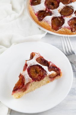 A piece of plum pie in a plate, a fork, a blurred image of the pie in the background.