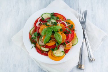 Salad of tomatoes and cucumbers with herbs in a light plate, fork, knife on a light table. Healthy food concept.