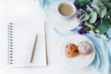 On the white table is a notepad, a pen, a cup of coffee, cupcakes in a plate, a bouquet of flowers.