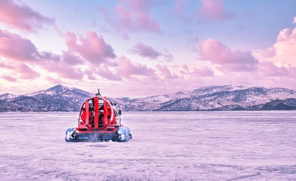 Hovercraft desliza rapidamente sobre a superfície do transparente congelado Lago Baikal ao pôr do sol, inverno paisagem rosa. — Fotografia de Stock