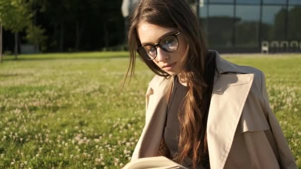 Stilvolles Mädchen mit Brille und braunem Umhang sitzt im Park auf dem Rasen und liest ein Buch. Schüler lernen. Geschenkpakete. Sonniger Sommerabend — Stockvideo