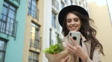 Kahverengi pelerinli ve siyah şapkalı güzel bir kadın elinde bir paket sebze, meyve ve elinde telefonla modern binaların yakınındaki caddede yürüyor. Yemek teslimatı