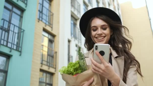 Piękna stylowa kobieta w brązowym płaszczu i czarny kapelusz idzie ulicą w pobliżu nowoczesnych budynków, trzymając w rękach paczkę warzyw i owoców oraz telefon. Dostawa żywności — Wideo stockowe