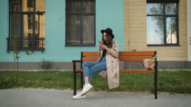 Portrét mladé podnikatelky v brýlích, pláštěnce a klobouku s upřímným úsměvem sedící na lavičce na pozadí moderní budovy držící telefon — Stock video