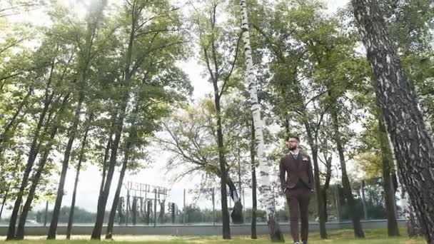 Brutalny biały mężczyzna z brodą w okularach i stylowym brązowym garniturze spaceruje po parku zastanawiając się nad biznesplanem — Wideo stockowe