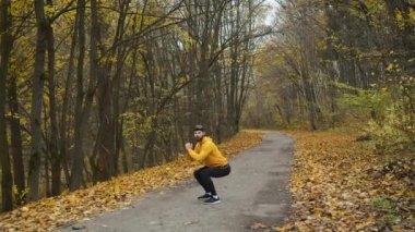 A bearded man of Caucasian appearance in yellow clothes warms up in the park and squats