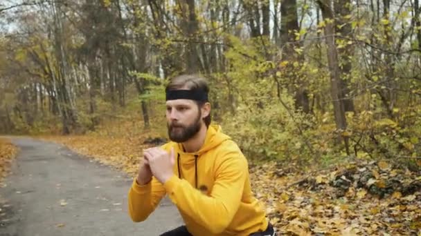 A bearded man of Caucasian appearance in yellow clothes warms up in the park and squats — Wideo stockowe