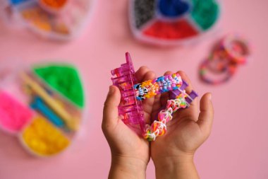 Lastik bantlardan yapılmış bir avuç örgülü bilezik, yapmak için bir makine ve bir çocuğun ellerinde dokumak için bir sapan. El motor becerilerinin geliştirilmesi için hobiler