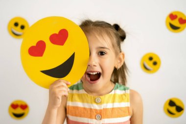Ağzı açık gülümseyen küçük bir kız, ellerinde gözleri yerine kalpleriyle karton bir gülümseme tutuyor, yüzünün bir kısmını onlarla örtüyor. Dünya emoji günü. Antropomorfik gülümseme.