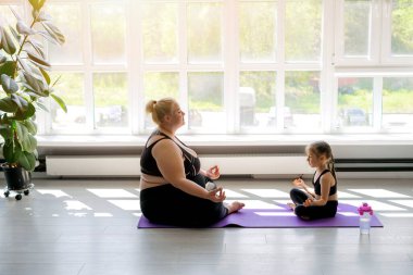 Şişman, tombul bir anne ve kırılgan, zayıf bir kız evde kocaman bir pencerenin yanında yoga yapıyorlar. Poz verip meditasyon yapan obez bir kadın ve bir çocuk. Kendini soyutlama için güneşli bir gün.