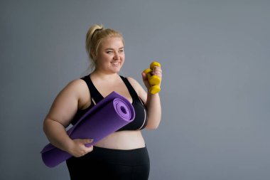 Şişko sarışın kadın pilates paspası ve sarı halterler tutuyor. Spor dersleri ve sağlıklı bir yaşam tarzı. Siyah spor giysili şişman kadın fotokopi alanından uzak duruyor. Mutlu model vücut pozitifi