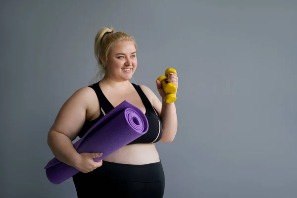 Fat Blonde woman holding pilates mat and yellow dumbbells. Fitness classes and a healthy lifestyle. Obese full-figured lady in black sportswear looks away copy space. Happy model body positive — Stock Image Fat Blonde Woman Holding Pilates Mat Yellow Dumbbells Fitness Classes Royalty Free Stock Images