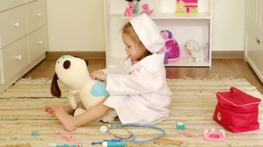 İki yaşındaki bir bebek doktorluk yapıyor. Kolunun altındaki peluş bir köpeğin termometresiyle vücut ısısını ölçüyor. Çocuk veteriner olmayı hayal ediyor.