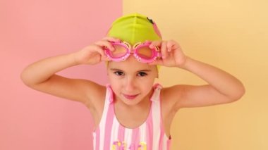 Mayo şapkalı ve çizgili pembe mayo giyen bir kız yengeç şeklinde komik çocuk yüzme gözlükleri takıyor. Sarı bir stüdyoda yüzücüyüm.