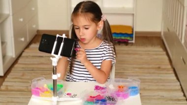 Masadaki bebek odasında, blogu için kamera telefonundan lastik bantlardan örgü örerek video kaydeden güzel bir kız. Küçük çocuk blogcu. Eğlenceli bir eğlence.